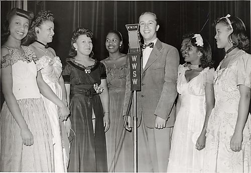 Man at WWSW radio station microphone flanked by seven young women in evening dress