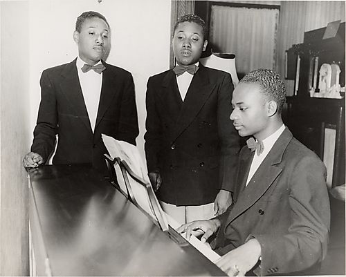 Three young men, one playing piano and two singing, in domestic interior