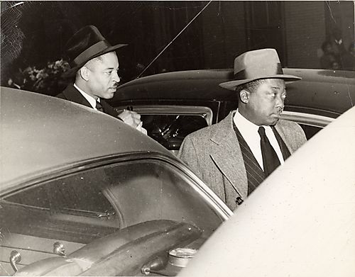 Two well-dressed men standing among parked cars