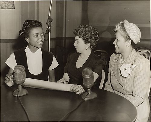 Three women, Ethel Ramos Harris, a singer from McKeesport, and a music teacher, at table with two microphones