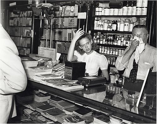 Penn Camera and Photo Supply Shop with woman fixing hair and man (Henry C. Cohn?) blowing nose behind counter