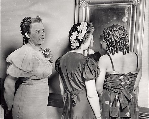 Three women in evening dresses and elaborate hair styles facing a mirror