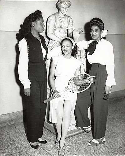 Three women, one holding a tennis racket, before a statue at Schenley High School