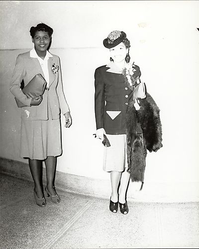Two women in suits, one holding handbag and fur, possibly for fashion show