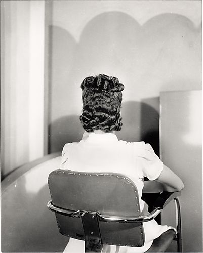 Woman wearing short sleeved white dress, in chair with back to camera modeling hair style, in studio