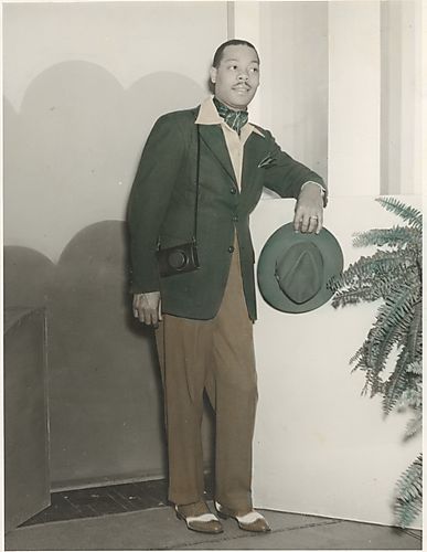 Fashion model Louie Smith with camera and hat standing near ferns