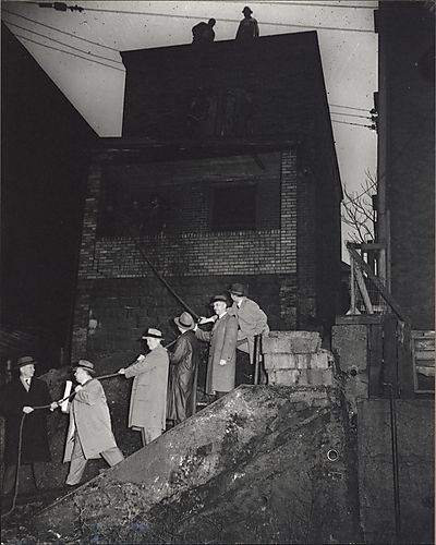 Six men on steps pulling on rope attached to pillar of porch of dilapidated house, with construction workers on porch roof and top-floor roof