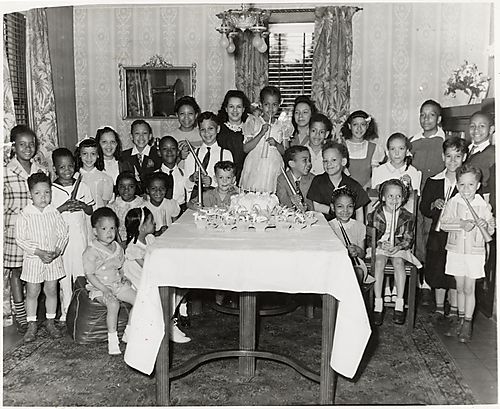 Group of children, some holding horns, gathered around table with cake and party favors at birthday party