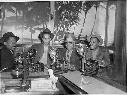 Copy of photograph of four photographers, including Paul Jones second from right and Walt Allen sitting on right end, at Crawford Grill No. 2 bar with cameras, tropical murals in background