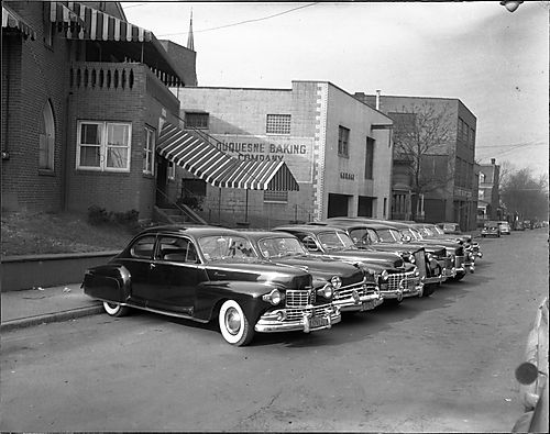 Row of cars parked in front of Gaines Funeral Home with garage with Duquesne Baking Company sign in background
